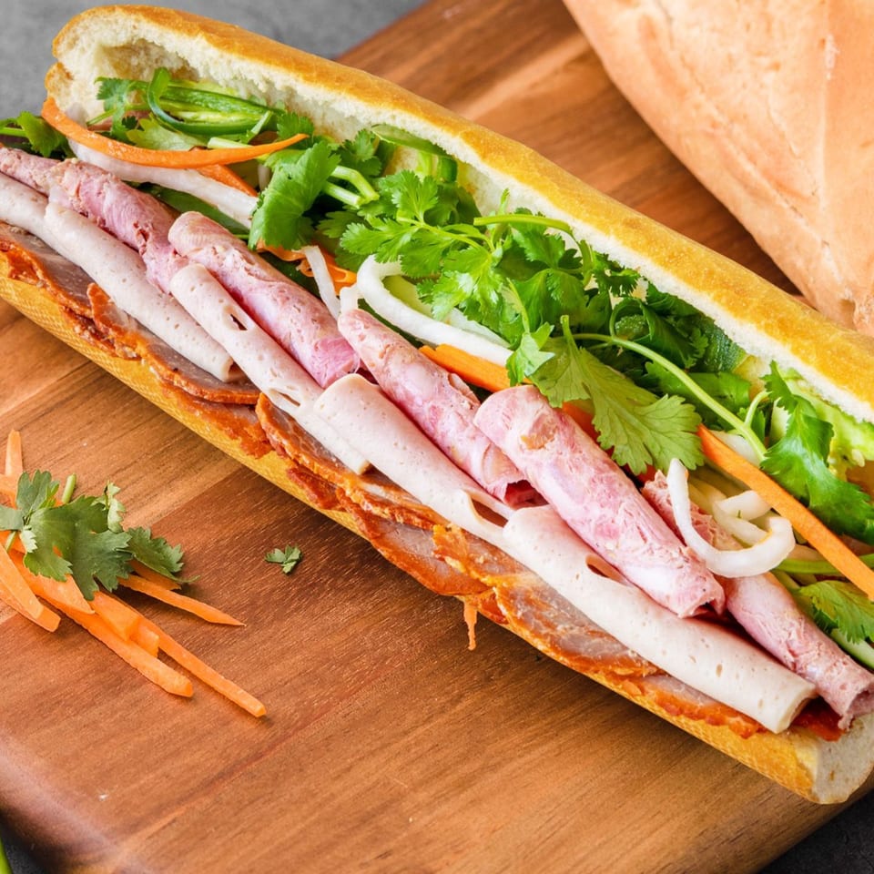 A Vietnamese banh mi sandwich, filled with various meats, fresh cilantro, carrots, and onions, on a wooden board.