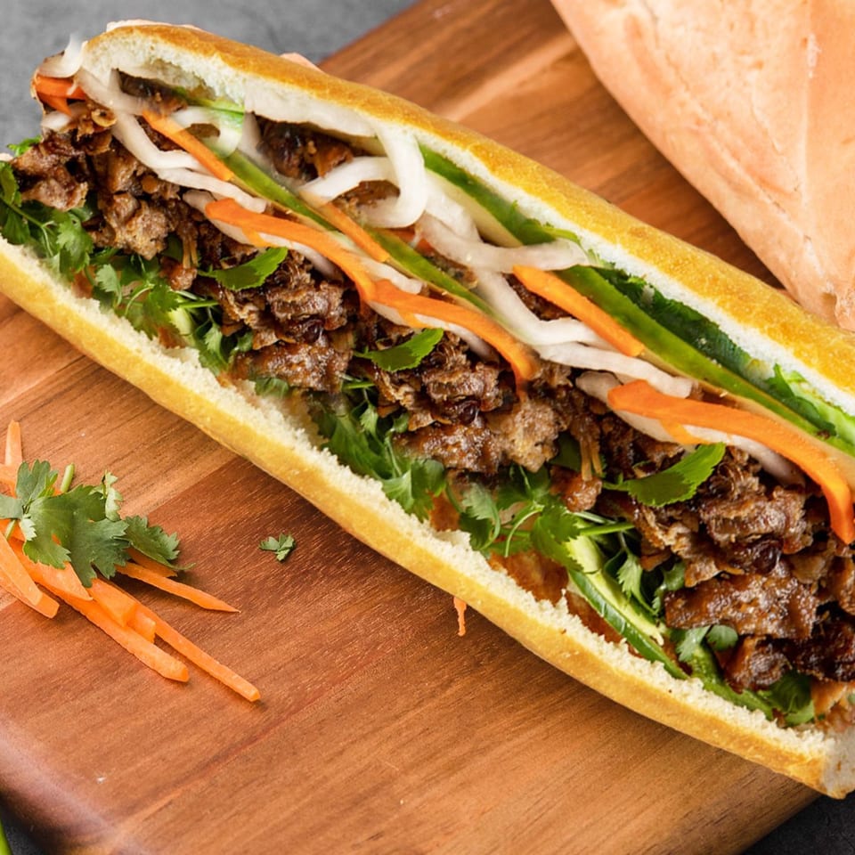 A Vietnamese banh mi sandwich with grilled pork, cilantro, pickled carrots, and daikon on a wooden board.